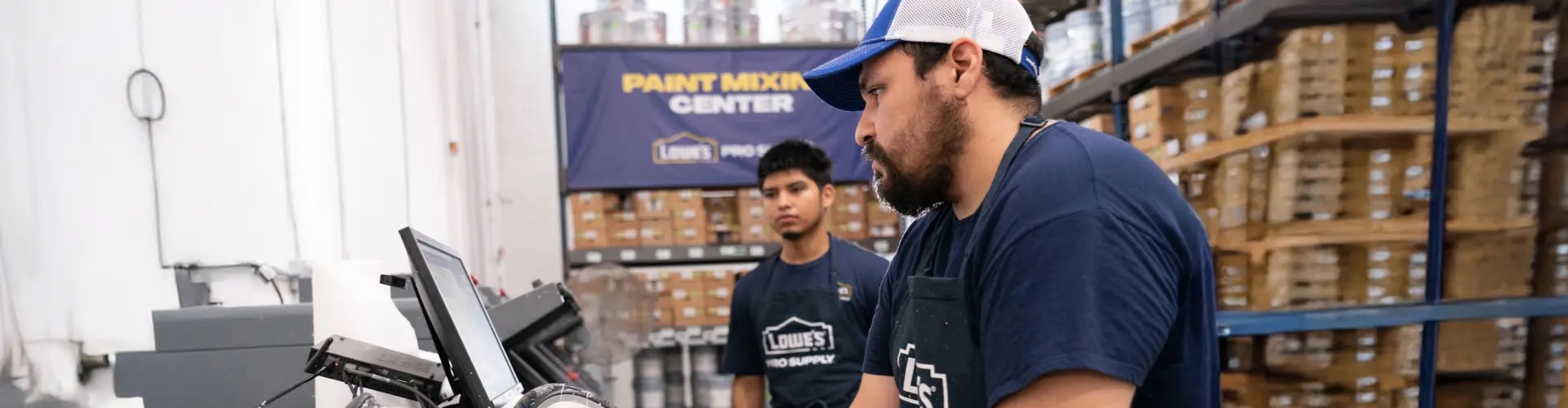 Two Lowe's associates in store uniform working at a paint-mixing centre terminal, with warehouse racking behind them.
