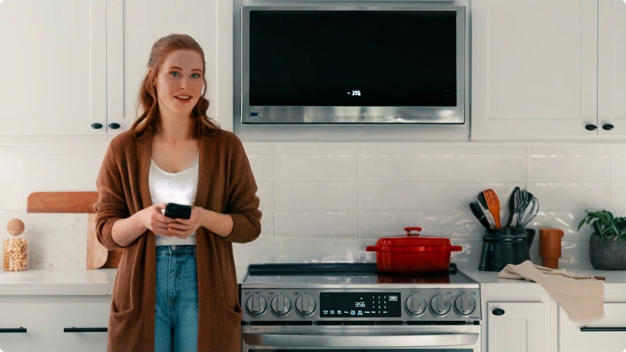A homeowner stands at her range with her phone in her hand, the microwave and oven clocks both reading 3:56.