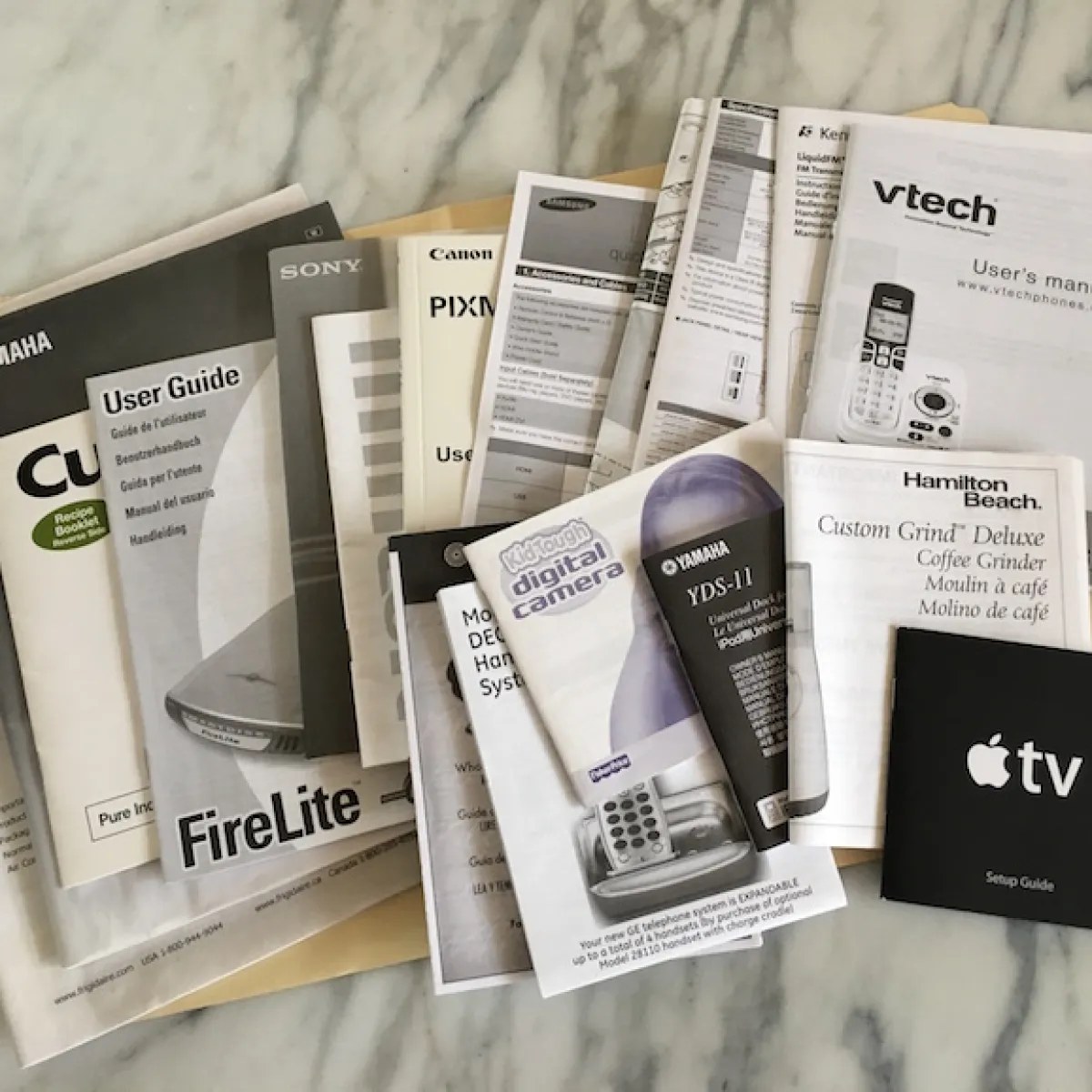A loose pile of printed appliance and electronics manuals spread across a marble counter — Yamaha, Sony, Canon, Samsung, vtech, Hamilton Beach and more.