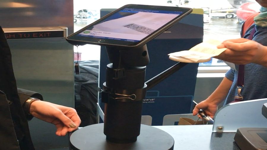 A passenger scans a paper boarding pass at the stand while the agent watches the iPad.